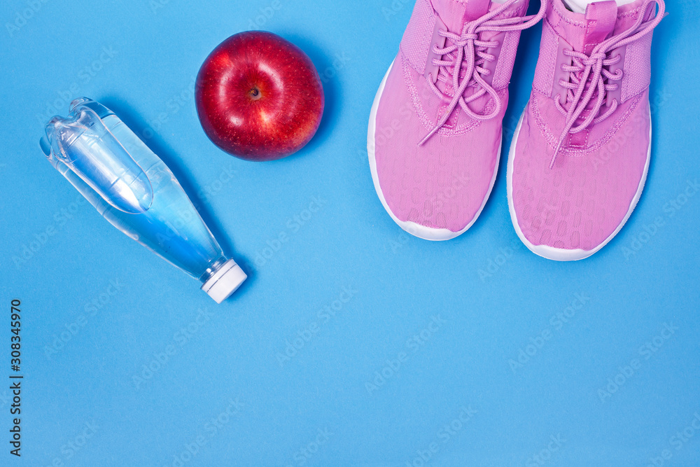 Fototapeta premium Pink sport shoes with red apple and bottle of water on a blue background. Concept healthy lifestyle, sport and diet. 