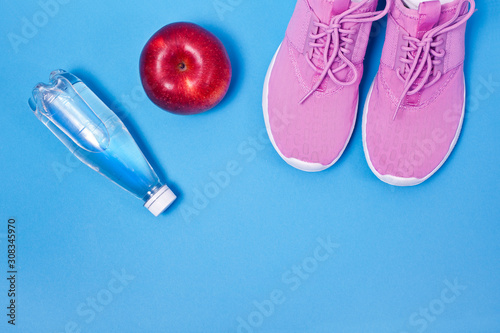 Pink sport shoes with red apple and bottle of water on a blue background. Concept healthy lifestyle, sport and diet. 