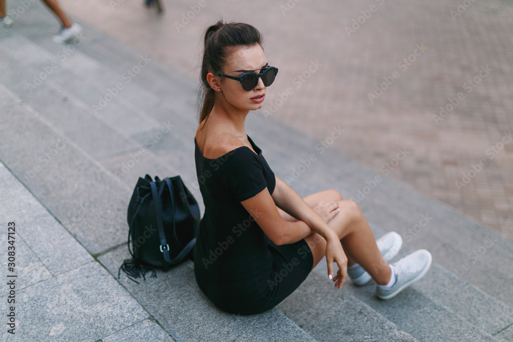Fototapeta premium Lonely girl with frown face, sits on the stairs in the city, in a black dress with round sunglasses, looking after her shoulder Full length portrait.