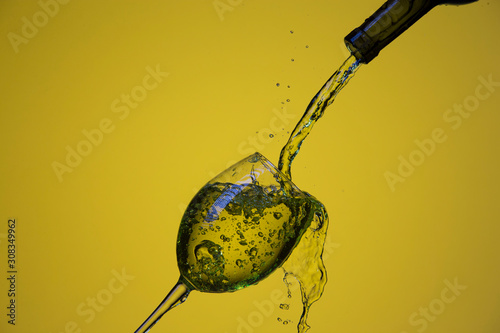 Drink Pouring Out of Bottle On The Right Hand Side to Wineglass with Colorful Liquid and Droplets. Over Lemon Color Background.