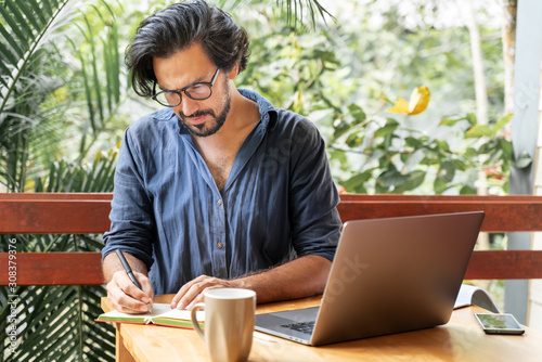 Handsome young curly latin man with glasses working on a laptop on the terrace of his house. Creative remote work, programmer, copywriter, writer