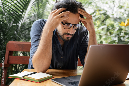 Handsome young curly latin man looks seriously intently at the laptop screen, holds his hands behind his head. Serious work, digital nomad, hard crisis, workaholism concept