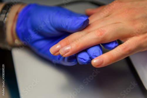 Professional manicure in salon, applying the base under the varnish. Hands of master in gloves and fingers of client, glare on nails, close-up.