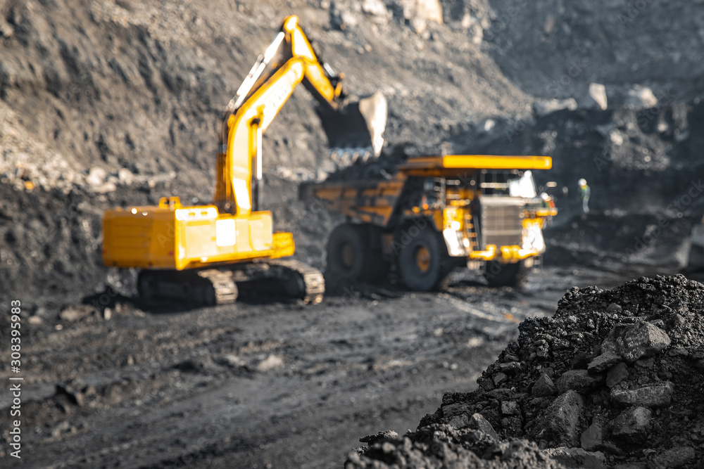 Focus on hard black coal, industrial quarry open mine. In background blurred loading of minerals anthracite excavator into large yellow truck