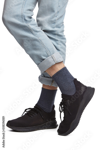 Cropped shot of a man's cross-legs in blue jeans and black sneakers, staying on a white background. It is dark grey socks on his foots. 
