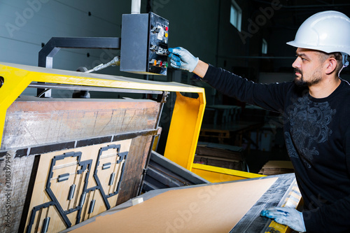 Worker in a hard hat turning on a punching machine. Cardboard boxes factory. Paper die cutting machine photo