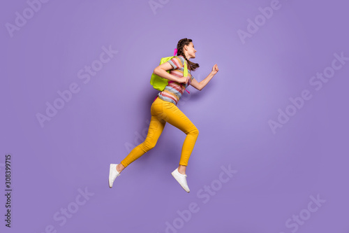 Full length profile photo of funky lady jumping high carry school bag rushing home after classes wear casual striped t-shirt yellow trousers isolated purple color background