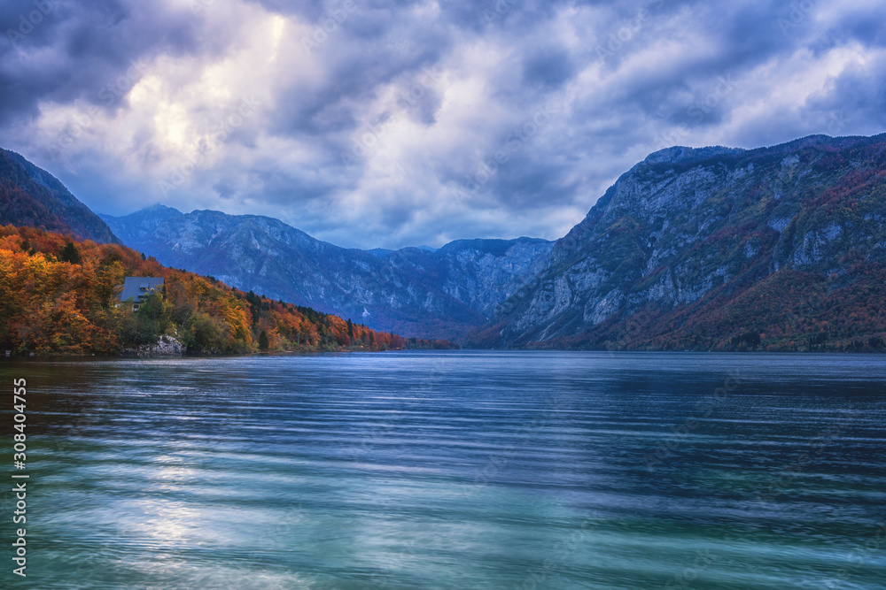 Bohinj lake (Bohinjsko jezero) in Triglav national park, Slovenia. Autumn landscape in the dusk, scenic view of the water, mountains, colored trees and cloudy sky, travel background of famous landmark Bohinj lake (Bohinjsko jezero) in Triglav national park, Slovenia. Autumn landscape in the dusk, scenic view of the water, mountains, colored trees and cloudy sky, travel background of famous landmark