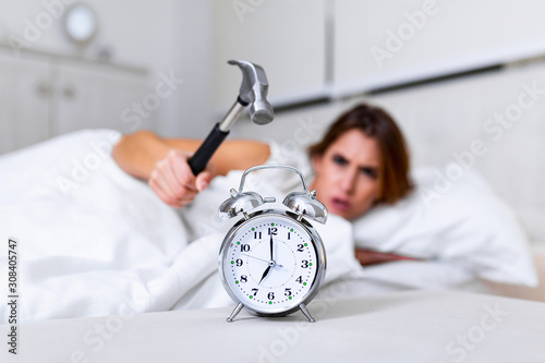 Young woman tries to break the alarm clock with hammer, Destroy the Clock. Girl lying in bed turning off an alarm clock with hammer in the morning at 7am. photo