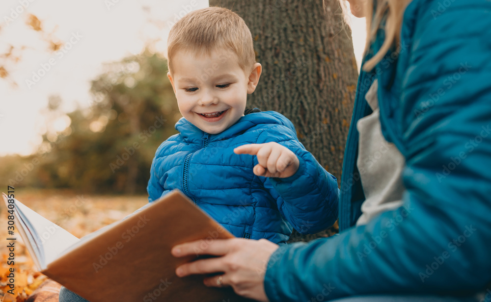 Fototapeta premium Portrait of a cute little boy reading a book outdoor with his mother smiling.