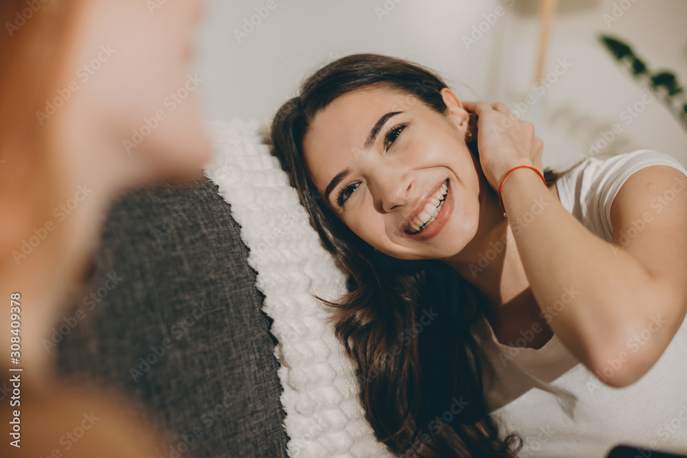 Fototapeta premium Portrait of a beautiful young woman laughing while leaning head on a bed touching her neck with the head looking to her friend.