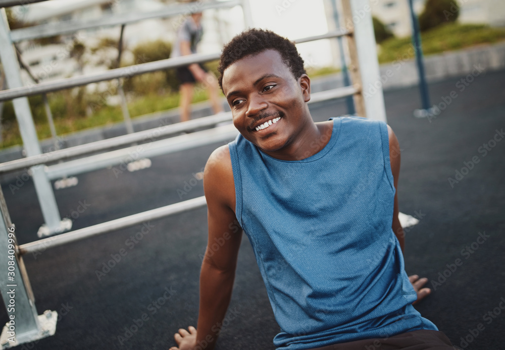 Cheerful fitness young sportsman resting after training at the outdoor gym park sitting on the ground smiling and looking away