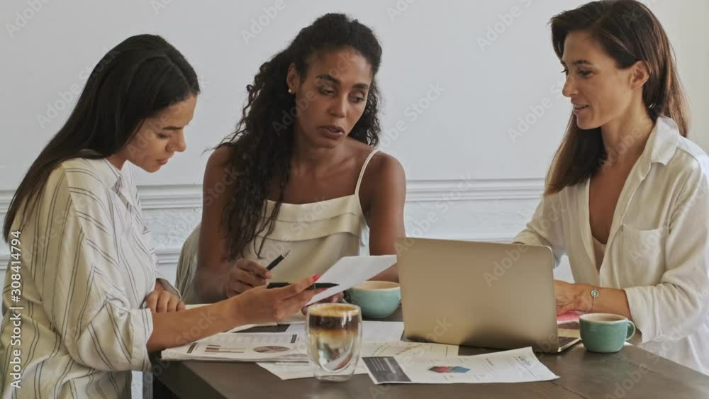 Three happy young beauty women colleagues working with documents and discussing while sitting by the table at office Three happy young beauty women colleagues working with documents and discussing while sitting by the table at office