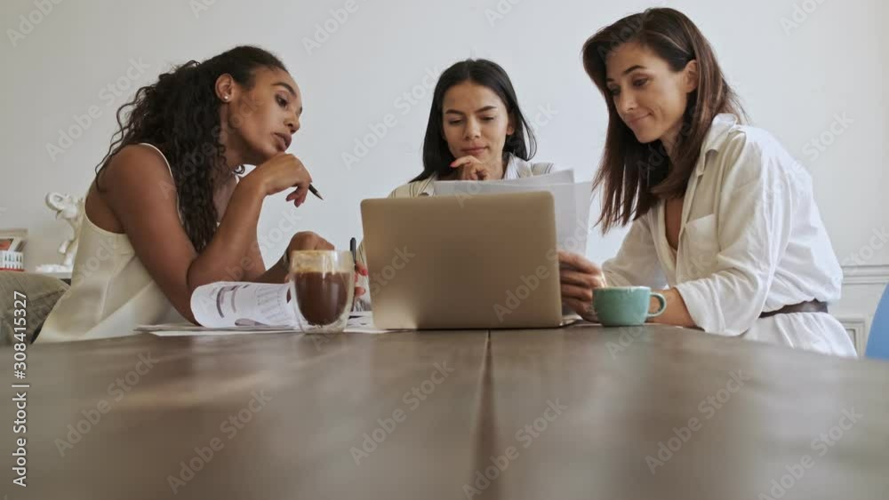 Three pleased young beauty women colleagues working with documents and laptop computer while having fun and sitting by the table at office