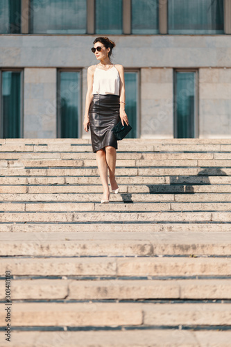Fashionable brunette businesswoman in leather pencil skirt and sunglasses walking on stairs