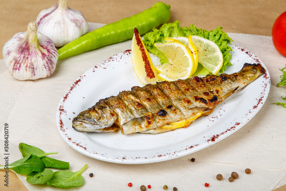 grilled trout with lemon and lettuce on a white plate. The plate is on white and gray wrapping paper, lined with pepper, peas, tomatoes, garlic, herbs