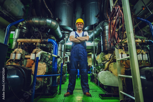 Low angle view of handsome caucasian worker in overall and helmet on head standing in ship next to engine with arms crossed. © dusanpetkovic1