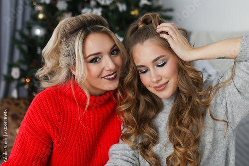 Two cute beautiful girls girlfriends sisters lovers in cozy bright red sweaters laugh smile and pose against the background of a Christmas tree in a decorated beautiful bright room.