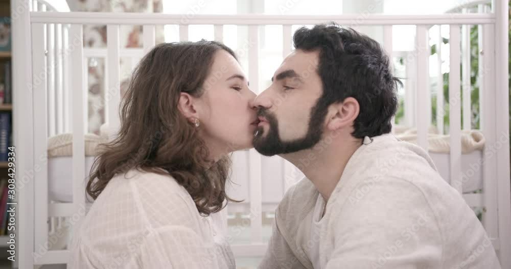 A Kiss and Gentle Embraces of a Man and a Woman. Young Family Expecting a Baby. Baby Cot on the Background. Concept of Parenthood, Happiness and Tenderness. A Kiss and Gentle Embraces of a Man and a Woman. Young Family Expecting a Baby. Baby Cot on the Background. Concept of Parenthood, Happiness and Tenderness.