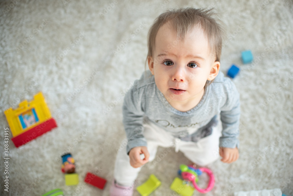 Portrait of one year old baby sitting on carpet in bright room looking up towards camera. Copy space on left. Portrait of one year old baby sitting on carpet in bright room looking up towards camera. Copy space on left.