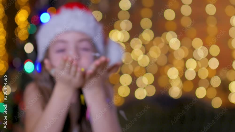 Little girl in santa claus hat blows and inflates small paper sweets from her hands. On a bright festive background. Little girl in santa claus hat blows and inflates small paper sweets from her hands. On a bright festive background.