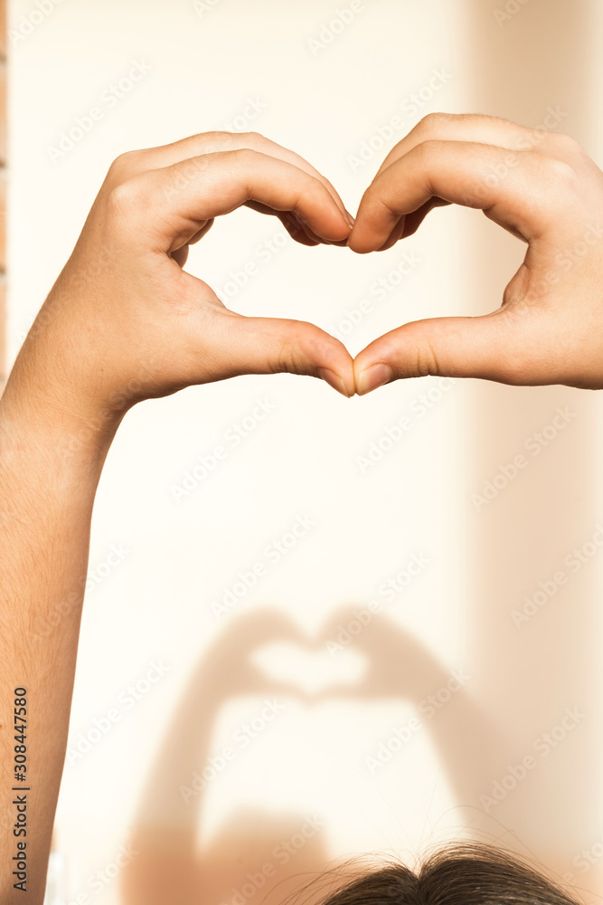 Obraz premium Hands of a teenage girl folded in the shape of a heart and a shadow from them on a light beige wall. The concept of Valentine's Day, love and friendship. Vertical walpaper mobile telephone composition