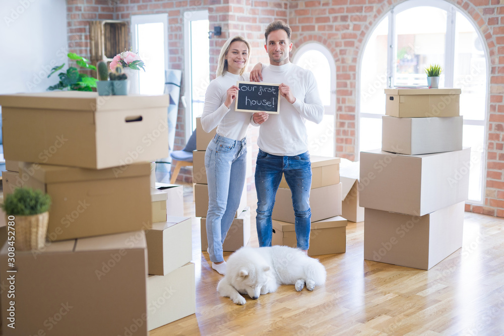 Fototapeta premium Young beautiful couple with dog standing holding blackboard with message at new home around cardboard boxes