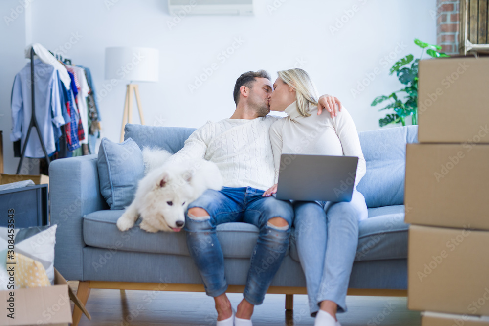Young beautiful couple with dog sitting on the sofa using laptop at new home around cardboard boxes