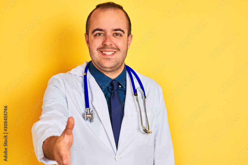 Young doctor man wearing coat and stethoscope standing over isolated yellow background smiling friendly offering handshake as greeting and welcoming. Successful business. Young doctor man wearing coat and stethoscope standing over isolated yellow background smiling friendly offering handshake as greeting and welcoming. Successful business.