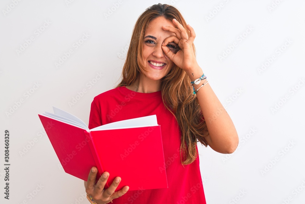 Young beautiful redhead woman reading a red book over isolated background with happy face smiling doing ok sign with hand on eye looking through fingers Young beautiful redhead woman reading a red book over isolated background with happy face smiling doing ok sign with hand on eye looking through fingers