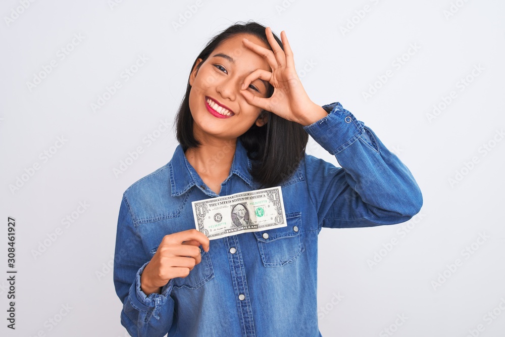 Young beautiful chinese woman holding one dollar standing over isolated white background with happy face smiling doing ok sign with hand on eye looking through fingers Young beautiful chinese woman holding one dollar standing over isolated white background with happy face smiling doing ok sign with hand on eye looking through fingers