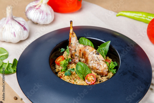 Fried rabbit leg with rice on a round black plate. Garnish is decorated with greens and tomatoes.