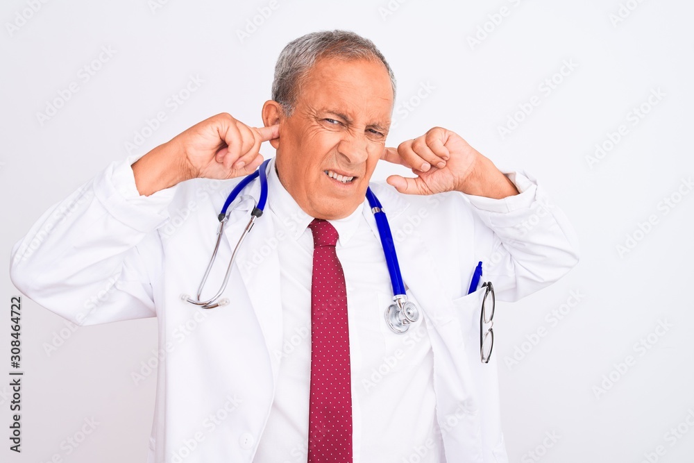 Senior grey-haired doctor man wearing stethoscope standing over isolated white background covering ears with fingers with annoyed expression for the noise of loud music. Deaf concept.