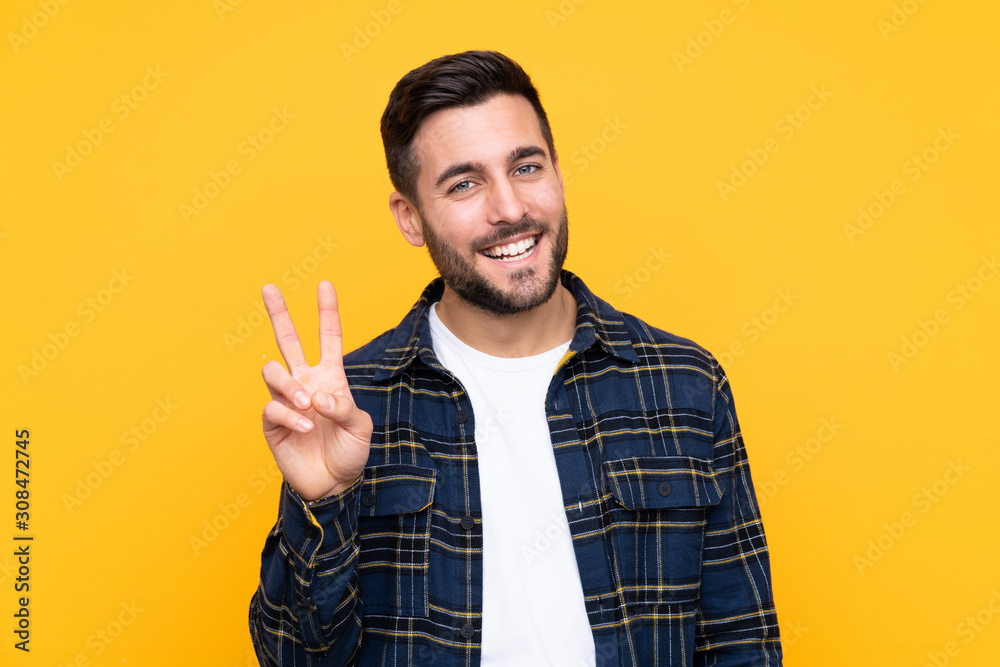 Young handsome man with beard over isolated yellow background smiling and showing victory sign