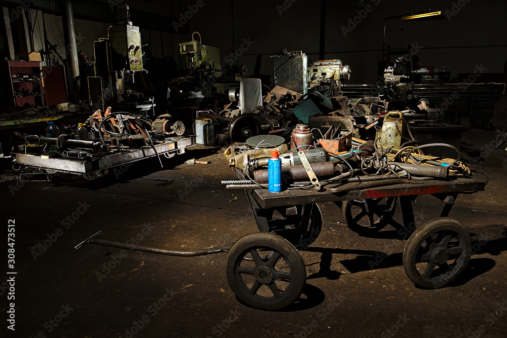 A dirty iron cart for manual movement stands in a dark workshop with tools. Dark cluttered workshop.