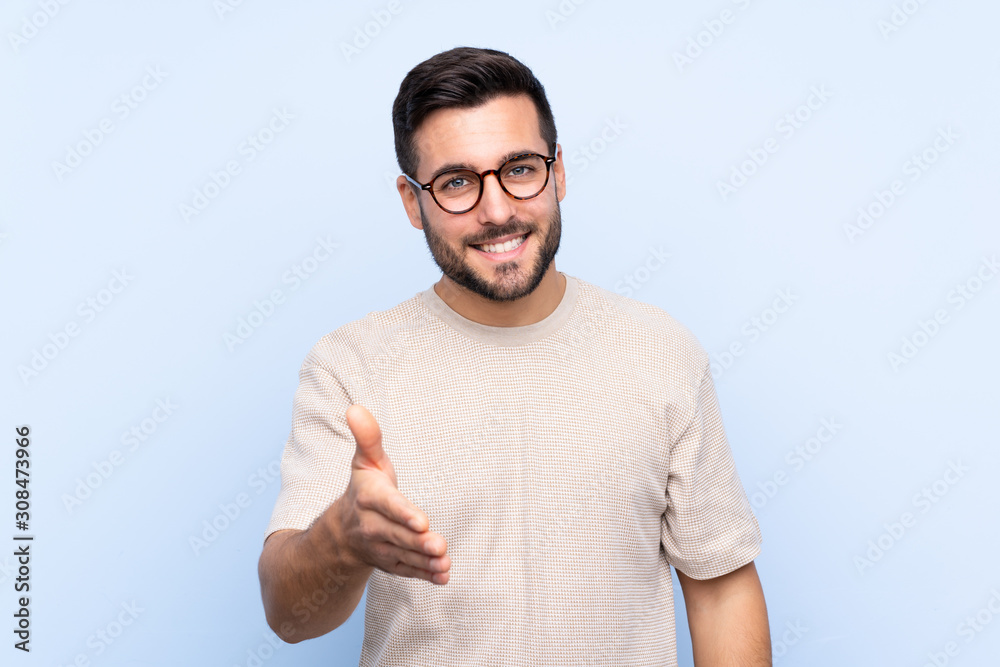 Young handsome man with beard over isolated blue background handshaking after good deal Young handsome man with beard over isolated blue background handshaking after good deal