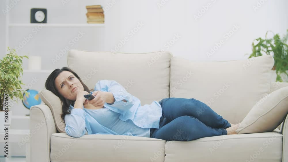 Young, relaxed woman is switching channels with zapper. She is watching TV, laying on sofa at home. Copy space. 4K. Slow motion. Young, relaxed woman is switching channels with zapper. She is watching TV, laying on sofa at home. Copy space. 4K. Slow motion.