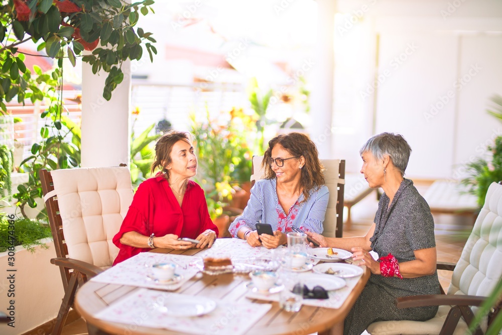 Meeting of middle age women having lunch and drinking coffee. Mature friends smiling happy using smartphone at home on a sunny day Meeting of middle age women having lunch and drinking coffee. Mature friends smiling happy using smartphone at home on a sunny day
