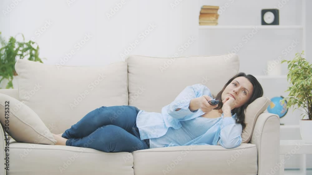 Young, smiling woman is switching channels with zapper. She is watching TV, laying on sofa at home. Copy space. 4K. Slow motion. Young, smiling woman is switching channels with zapper. She is watching TV, laying on sofa at home. Copy space. 4K. Slow motion.