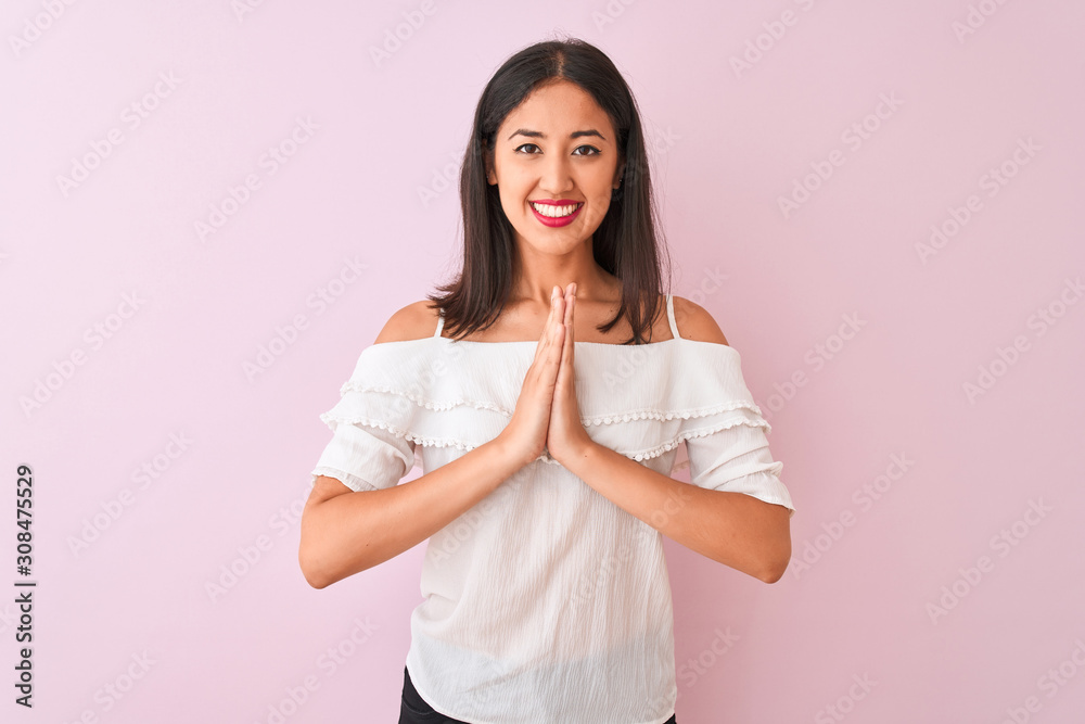 Beautiful chinese woman wearing white t-shirt standing over isolated pink background praying with hands together asking for forgiveness smiling confident.