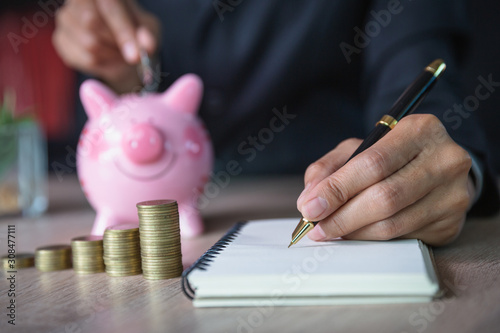 The hand of a business person is going to put a coin in a pink piggy bank. Hand was about to write an income and expense account.Financial accounting concepts. Calculate assets for business success.