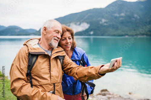 A senior pensioner couple standing by lake in nature, taking selfie.