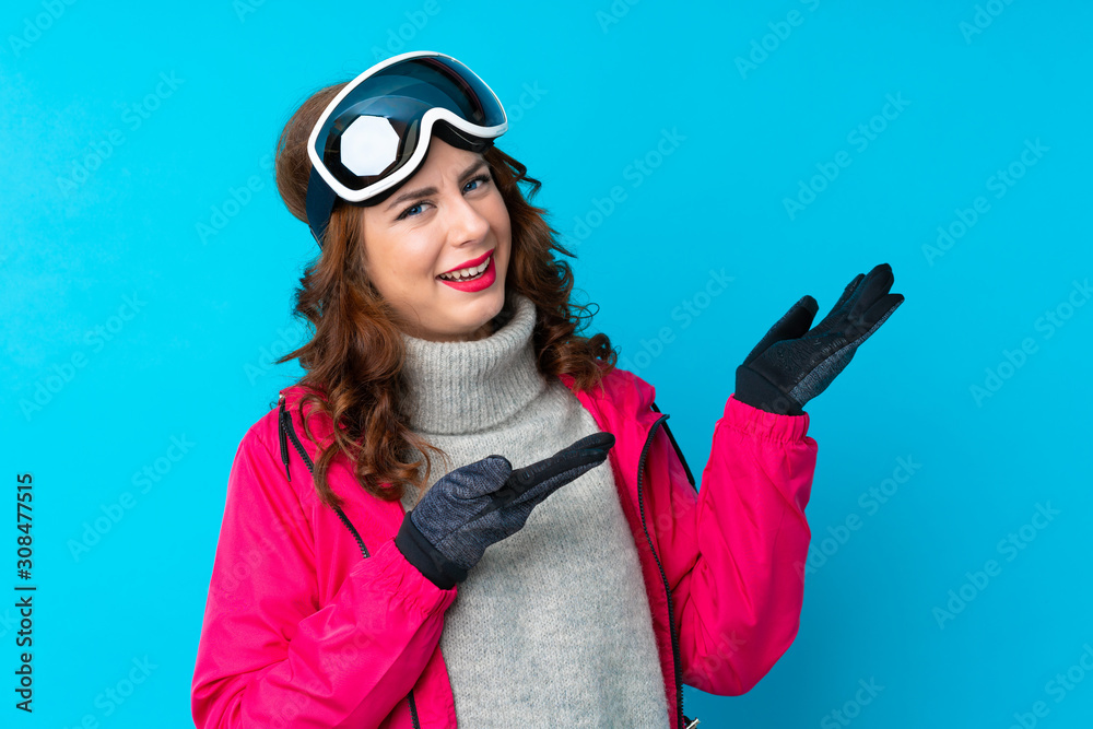 Skier woman with snowboarding glasses over isolated blue wall extending hands to the side for inviting to come