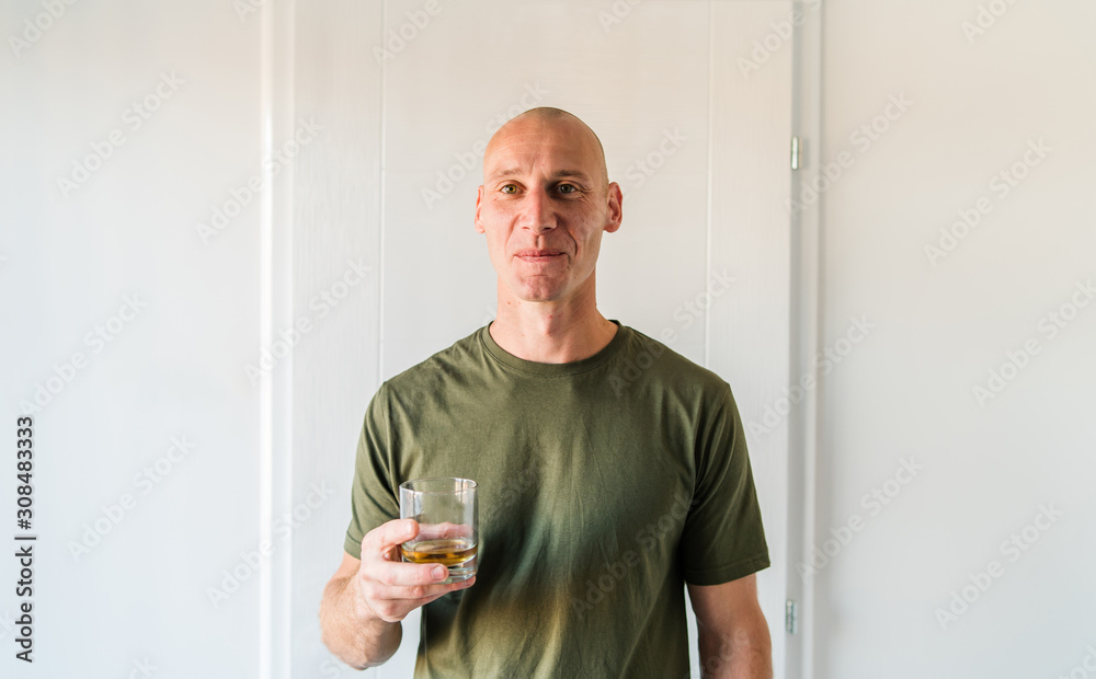 Portrait of young caucasian man good looking with short hair wearing green  t shirt holding a glass of brandy or whiskey alcohol drink standing in  front of white wall at home Stock