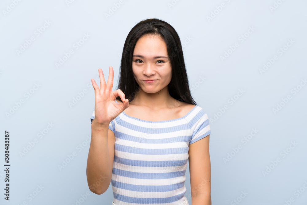 Teenager asian girl over isolated blue background showing an ok sign with fingers Teenager asian girl over isolated blue background showing an ok sign with fingers