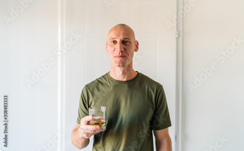 Portrait of young caucasian man good looking with short hair wearing green t shirt holding a glass of brandy or whiskey alcohol drink standing in front of white wall at home