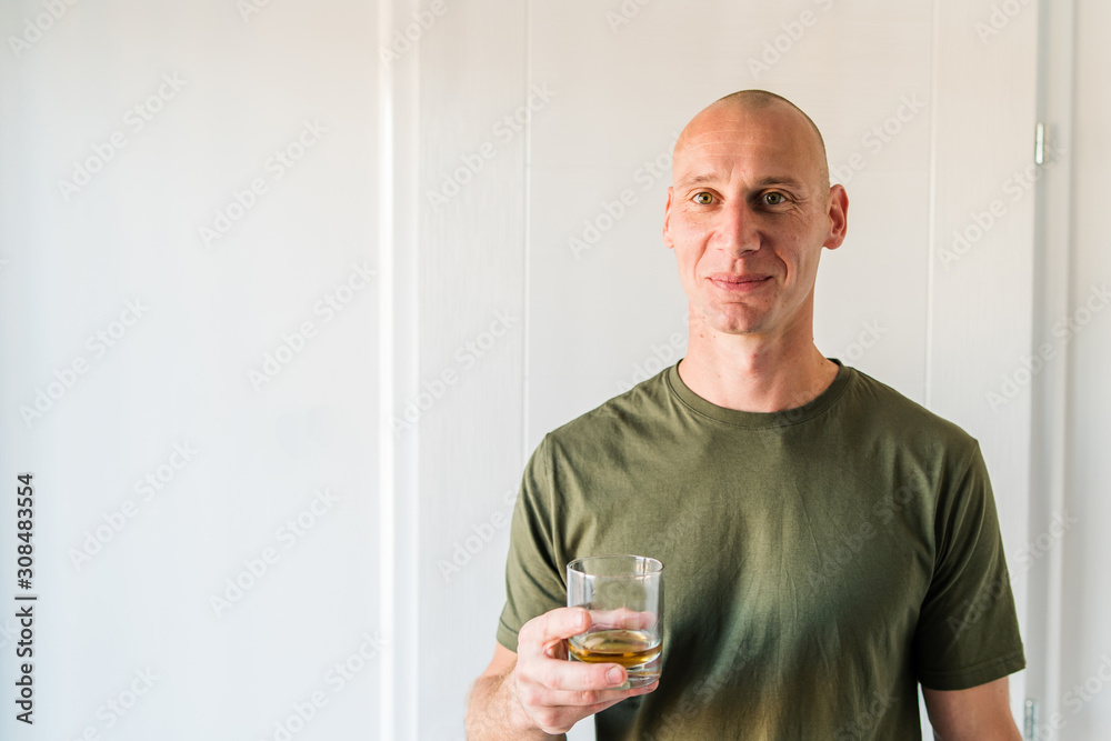 Portrait of young caucasian man good looking with short hair wearing green t shirt holding a glass of brandy or whiskey alcohol drink standing in front of white wall at home Portrait of young caucasian man good looking with short hair wearing green t shirt holding a glass of brandy or whiskey alcohol drink standing in front of white wall at home