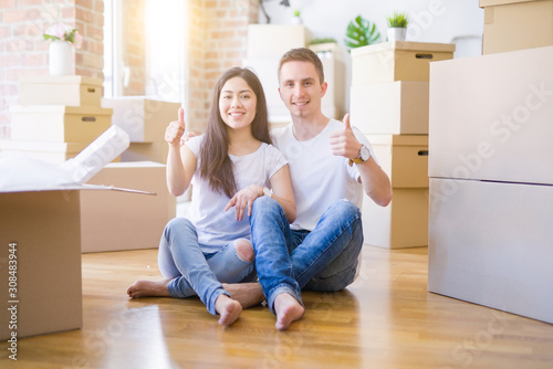 Young beautiful couple sitting on the floor at new home around cardboard boxes happy with big smile doing ok sign, thumb up with fingers, excellent sign