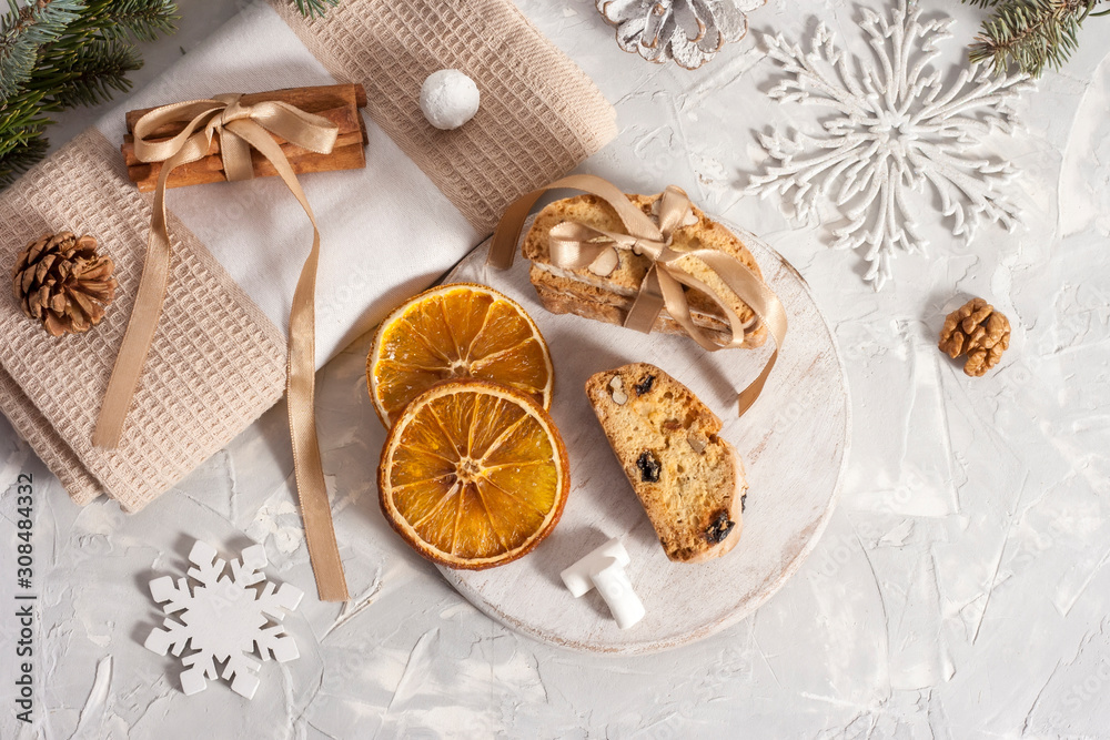 Obraz premium Traditional Italian biscotti or cantuccini cookies on a round white wooden board with cinnamon, dried orange slices and marshmallows on a light table near a Christmas tree. Christmas baking concept