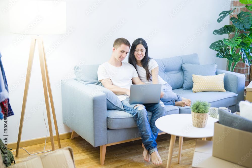 Young beautiful couple sitting on the sofa using laptop at new home around cardboard boxes Young beautiful couple sitting on the sofa using laptop at new home around cardboard boxes